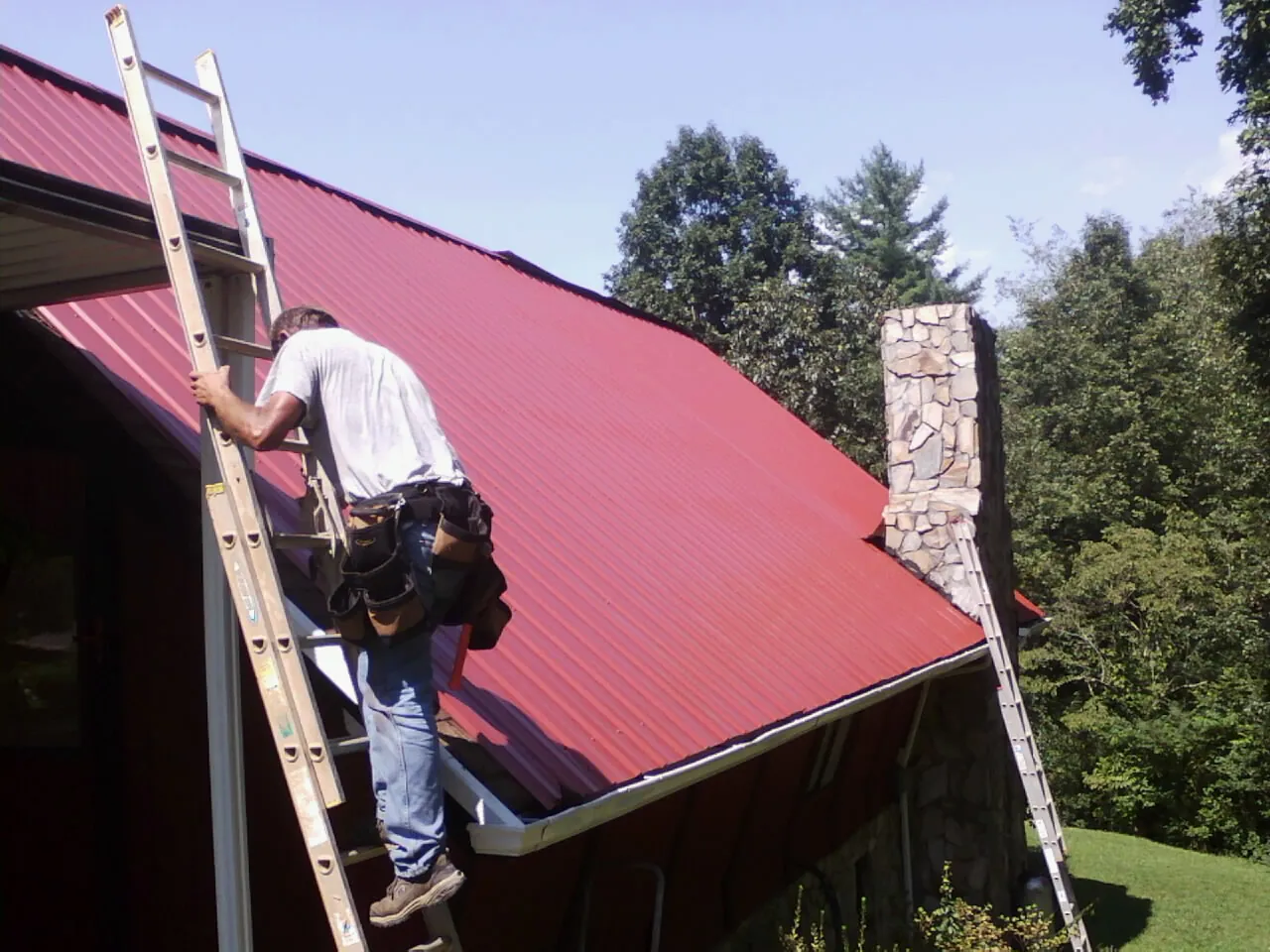 Roofer on a ladder beside a red metal roof and stone chimney — Soffit & Fascia Repair by Roofing Brothers in Haralson